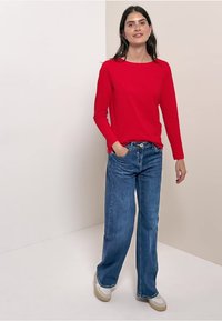 Chemise rouge à manches longues associée à un jean large bleu et des baskets beiges, le tout sur un fond neutre. Tenue simple et décontractée.