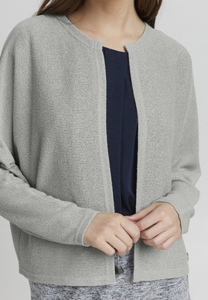 Femme portant un cardigan en maille gris clair ouvert sur le devant, par-dessus un haut bleu foncé avec un pantalon gris à motifs, les mains délicatement jointes à la taille.