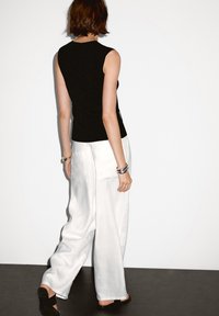 Donna che indossa un top senza maniche nero, pantaloni bianchi larghi, sandali con tacco neri e braccialetti d'argento, in piedi contro un muro bianco semplice.