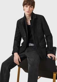Zwarte suède jas met een getailleerde pasvorm, voorzien van een hoge kraag, knoopsluiting en contrasterende stiksels. G gedragen over een zwarte top en grijze jeans.
