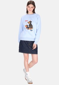 Lichtblauwe sweatshirt met een hondenafbeelding op de voorkant, gecombineerd met een donker denim rokje. Model draagt zilveren sneakers en witte sokken.