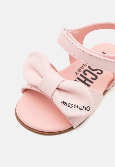 Rozā čības, kas izgatavotas no gludas ādas, ar lielu lentes detalizāciju un zīmola nosaukumu "Moschino" izšūtu malā. Atvērta dizaina.