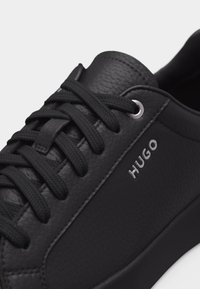 Baskets en cuir noir texturé avec lacets noirs et œillets argentés, arborant la marque "HUGO" sur le côté.