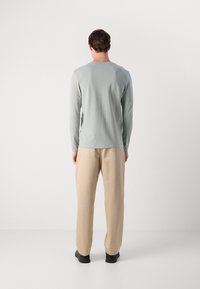 Mężczyzna stoi tyłem, ubrany w jasnoszary longsleeve, beżowe spodnie i czarne buty, na tle białego, gładkiego tła.