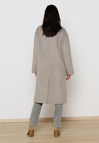 Lichtgrijze wollen blendjas met een losse pasvorm, lange mouwen en een naden in het midden van de rug. Gecombineerd met vervaagde grijze jeans en bruine haklaarzen.
