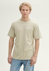 Lichtbeige katoenen T-shirt met korte mouwen en een ronde hals. Bevat rode en grijze tekst op de linkerkant van de borst. G gedragen met blauwe spijkerbroek.