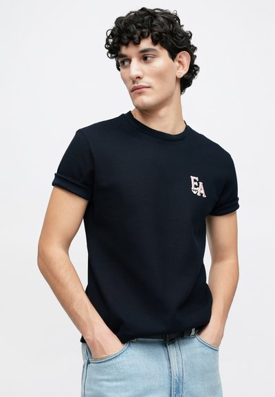 Μπλούζα με υφή σε ναυτικό μπλε χρώμα, με γυρισμένα μανίκια και μικρό ροζ κεντημένο λογότυπο "EA" στο αριστερό στήθος. Φορεμένη με ανοιχτόχρωμο μπλε τζιν.