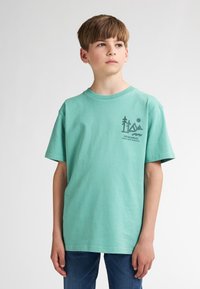 Tricou din bumbac verde mentă cu un design grafic pe partea stângă a pieptului. Tricoul are mâneci scurte și gât rotund. Jeanșii din denim completează ținuta.