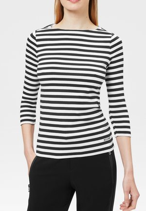 Femme portant un haut à rayures horizontales noires et blanches à manches longues 3/4 et un pantalon noir avec des poches zippées.