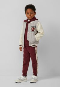 Grijs en crèmekleurig varsityjack met borduursels van "NUMBER" en "STUNT", maroon hoodie, bijpassende trainingsbroek met zijstrepen en witte sneakers.