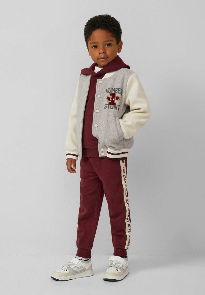 Grijs en crèmekleurig varsityjack met borduursels van "NUMBER" en "STUNT", maroon hoodie, bijpassende trainingsbroek met zijstrepen en witte sneakers.