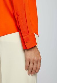 Ljus orange långärmad blus med en knappslå och knappdetaljer på manchetten, i kombination med krämfärgade byxor. Slät tygtextur.