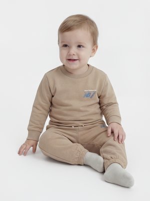 Criança pequena com cabelo castanho claro sentada, vestindo sweatshirt e calças bege da New Balance com meias claras, sorrindo ligeiramente.
