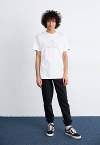 Weißes Baumwoll-T-Shirt mit blauer Bedruckung, kombiniert mit schwarzen, schmal zulaufenden Jogginghosen und schwarz-weißen Retro-Sneakers. Einfarbiger Hintergrund.
