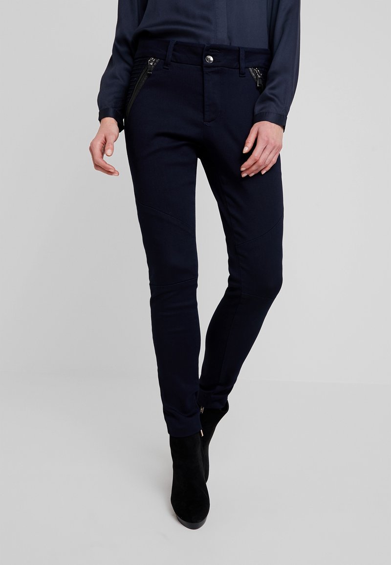 Kvinna klädd i slim-fit marinblå byxor med framlåst fickor och svarta ankelskor, stående mot en ljusgrå bakgrund.