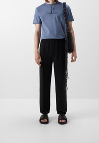 Maglietta in cotone blu con logo ricamato, pantaloni neri con vita elastica, borsa a tracolla intrecciata nera e sandali neri.