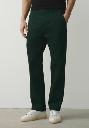 Egy férfi, aki sötétzöld egyenes szárú nadrágot és fehér tornacipőt visel, egy kézzel a zsebében áll, egyszínű világos háttér előtt.