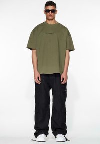 Olive green oversized t-shirt with black logo, paired with black cargo pants featuring multiple pockets, and white sneakers.