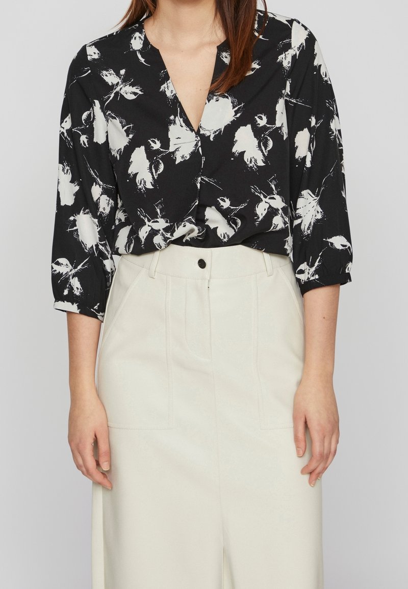 Blusa nera con fantasia floreale bianca, scollatura a V e maniche a tre quarti. Abbinata a pantaloni chiari con tasche frontali.