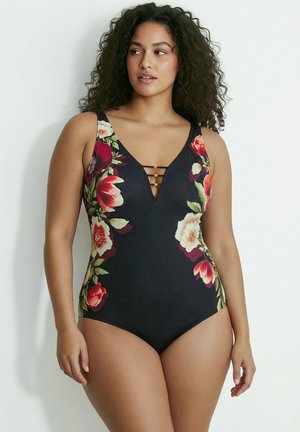 Femme grande taille aux cheveux bouclés portant un maillot de bain une pièce noir avec des motifs floraux colorés et des bretelles décoratives au niveau de l'encolure.