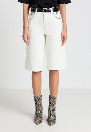 Frau trägt weiße knielange Shorts mit genietetem Taschen, schwarzem Gürtel und knöchelhohen Stiefeln im Schlangenledermuster, steht vor einem einfarbigen Hintergrund.