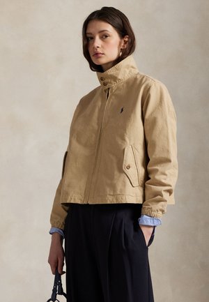 Vrouw draagt beige jas met rits en hoge kraag en donkerblauwe broek, houdt een kleine donkere handtas vast tegen een neutrale achtergrond.