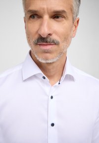 Mænds hvid skjorte med knapper og spids krave, med mørkeblå knapper og en glat tekstur. Nærbillede af overkroppen og ansigtet.