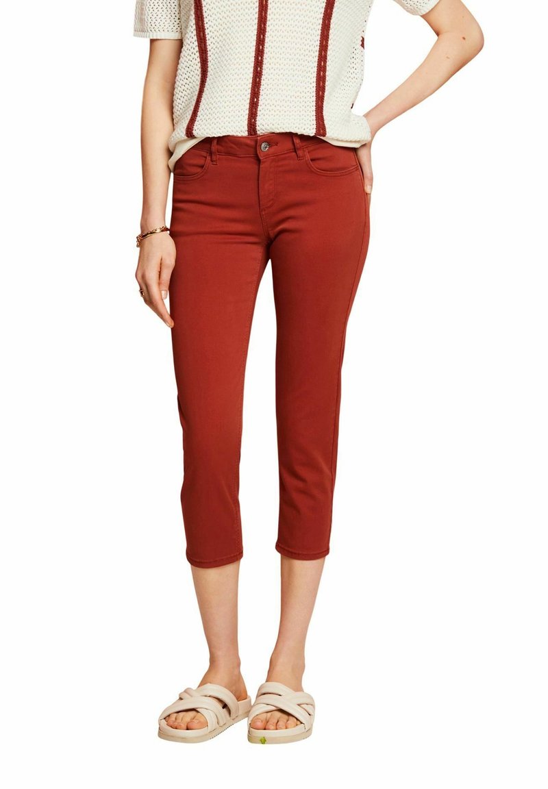 Personne portant un pantalon court couleur rouille, un haut en maille crème avec des rayures marron et des sandales beiges, debout avec une main sur la hanche.