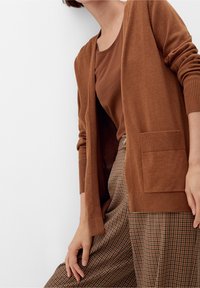 Hnědý kardigan z měkké látky, otevřený přední díl s žebrovanými manžetami a lemem, s dvěma předními kapsami; doplněno hnědým topem a károvanými kalhotami.