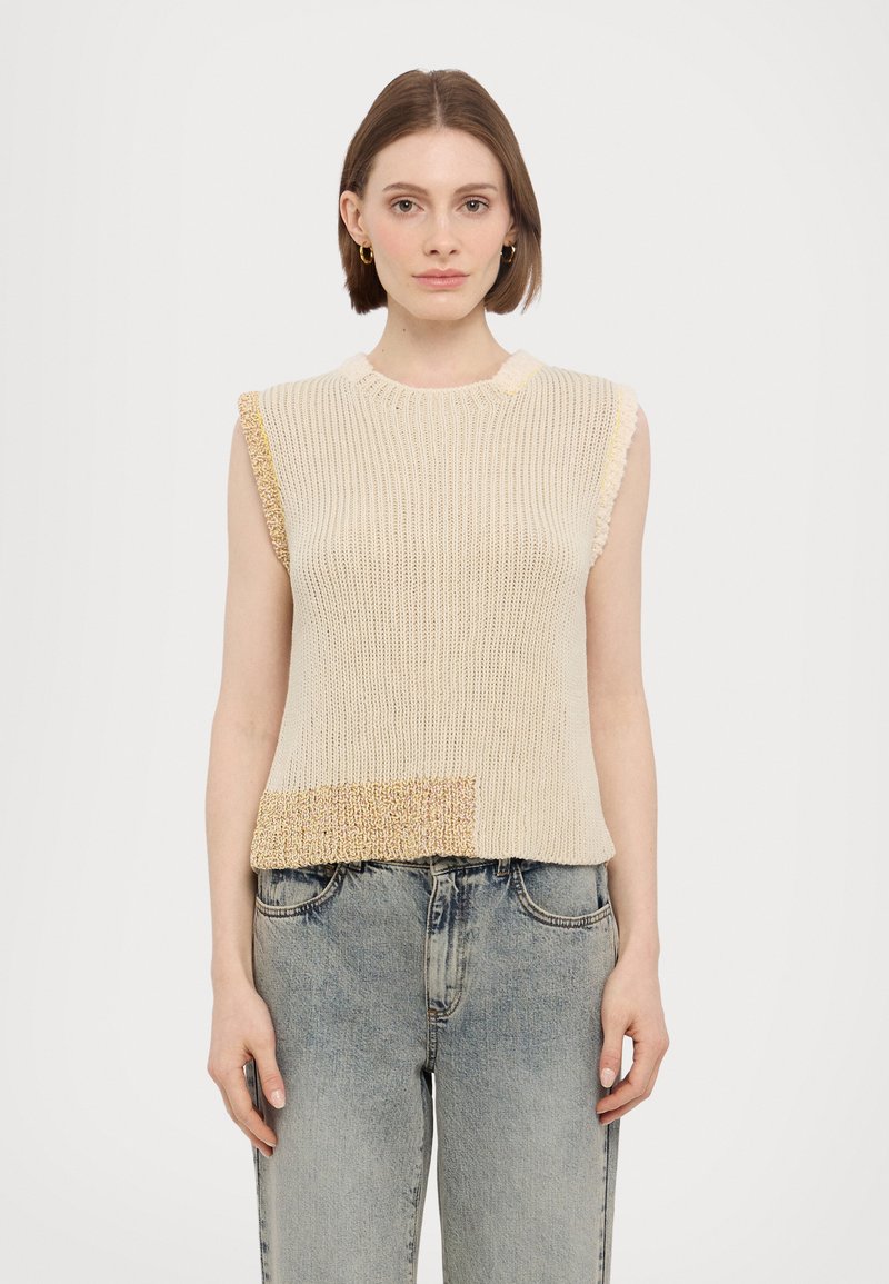 Beige gebreide mouwloze top met een gestructureerde afwerking en een met goud accentueerde patch aan de onderkant. Gecombineerd met lichtblauwe, licht versleten jeans.