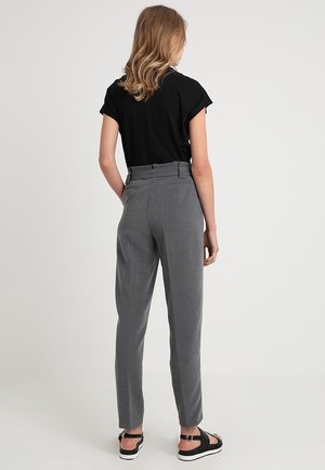 Femme aux cheveux blonds ondulés portant un haut noir à manches courtes, un pantalon gris taille haute et des sandales compensées noires, debout de dos.