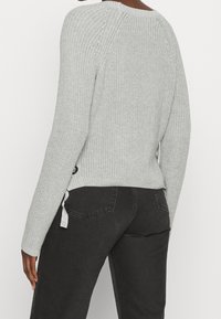 Szary sweter dziany o luźnym kroju, z prążkowaną fakturą, wiązaniami po bokach i opuszczonymi szwami na ramionach, zestawiony z czarnymi dżinsami.