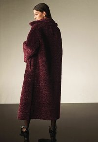 Lang, oversized frakke i dyb burgunder, lavet af et skridsikkert, blødt materiale. Har en bred krave og fulde ærmer, der når til anklerne.