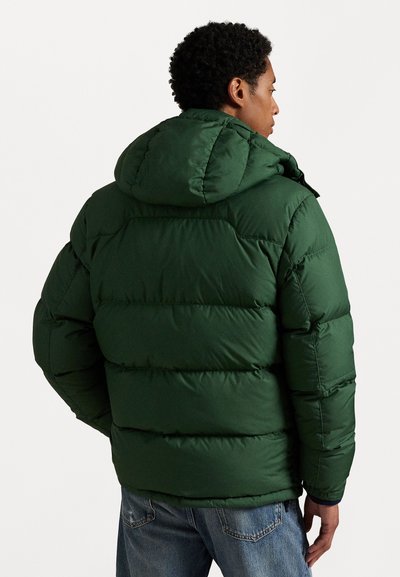 Grön pufferjacka med huva, med kviltade sektioner och en slät yta. Jackan bärs med blå denimjeans, vilket ger en avslappnad look.