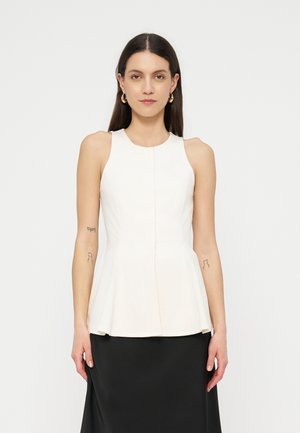 Kvinde med langt mørkt hår iført hvid ærmeløs peplum-top, sort nederdel, guldringøreringe og viser små tatoveringer på begge underarme.