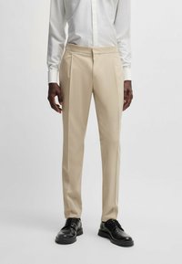 Beige Maßhose mit Falten, aus leichtem Stoff gefertigt. Kombiniert mit schwarzen Schnürschuhen und einem weißen Langarmhemd.
