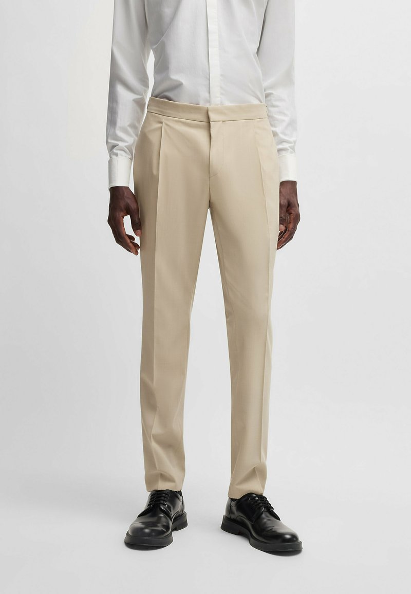 Beige Maßhose mit Falten, aus leichtem Stoff gefertigt. Kombiniert mit schwarzen Schnürschuhen und einem weißen Langarmhemd.