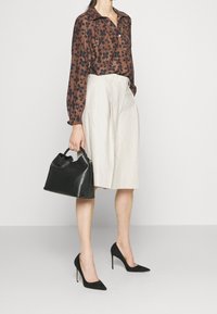 Blusa marrone con motivo floreale e colletto, gonna plissettata beige, scarpe con tacco nere e borsa nera strutturata.
