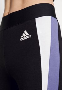 Czarne legginsy z szerokim pasem, trzema pionowymi paskami w kolorach fioletowym, białym i czarnym, z białym logo Adidas na lewym biodrze.