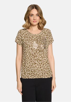 Kvinde med skulderlangt blondt hår iført en beige og brun leopardmønstret bluse med korte ærmer og sorte bukser, vendt fremad.