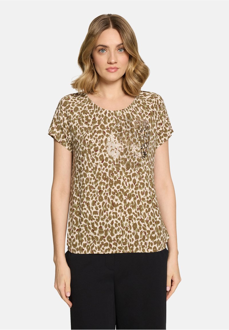Femme aux cheveux blonds mi-longs portant un haut à manches courtes beige et marron à motif léopard et un pantalon noir, regardant en avant.