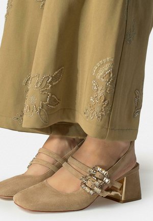 Zapatos bajos de ante beige con doble tira y hebillas adornadas con gemas, combinados con pantalones de pierna ancha bordados en marrón.