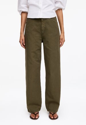 Personne portant un pantalon large vert olive, une chemise blanche boutonnée et des sandales marron foncé, debout devant un fond uni.