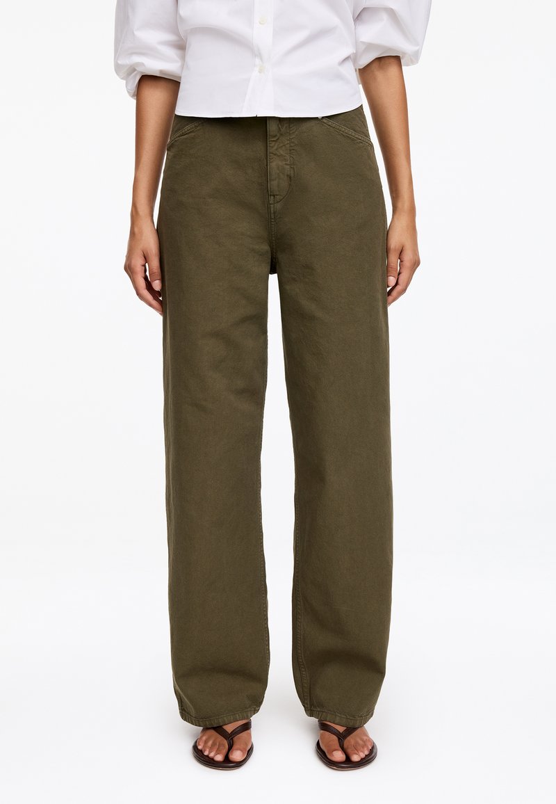 Personne portant un pantalon large vert olive, une chemise blanche boutonnée et des sandales marron foncé, debout devant un fond uni.