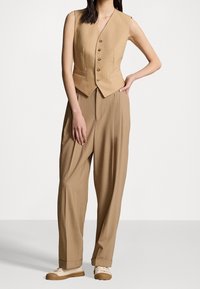 Béžový dvoudílný outfit s bezrukávovou, přiléhavou vestou na knoflíky a kalhotami s širokými nohavicemi. Hladká látka s na míru šitým střihem.