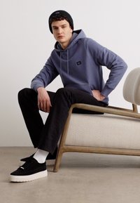 Felpa blu con tasca frontale, abbinata a pantaloni neri e sneakers nere. Il modello è seduto su una sedia di legno con un cuscino testurizzato.
