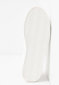 Semelle de chaussure blanche avec un motif de bande de roulement en zigzag, positionnée verticalement contre un coin de mur et de sol blancs.