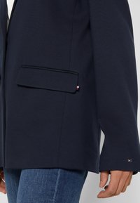 Personne portant un blazer bleu marine foncé avec une poche à rabat et un petit logo tricolore, associé à un jean en denim bleu, main détendue sur le côté.