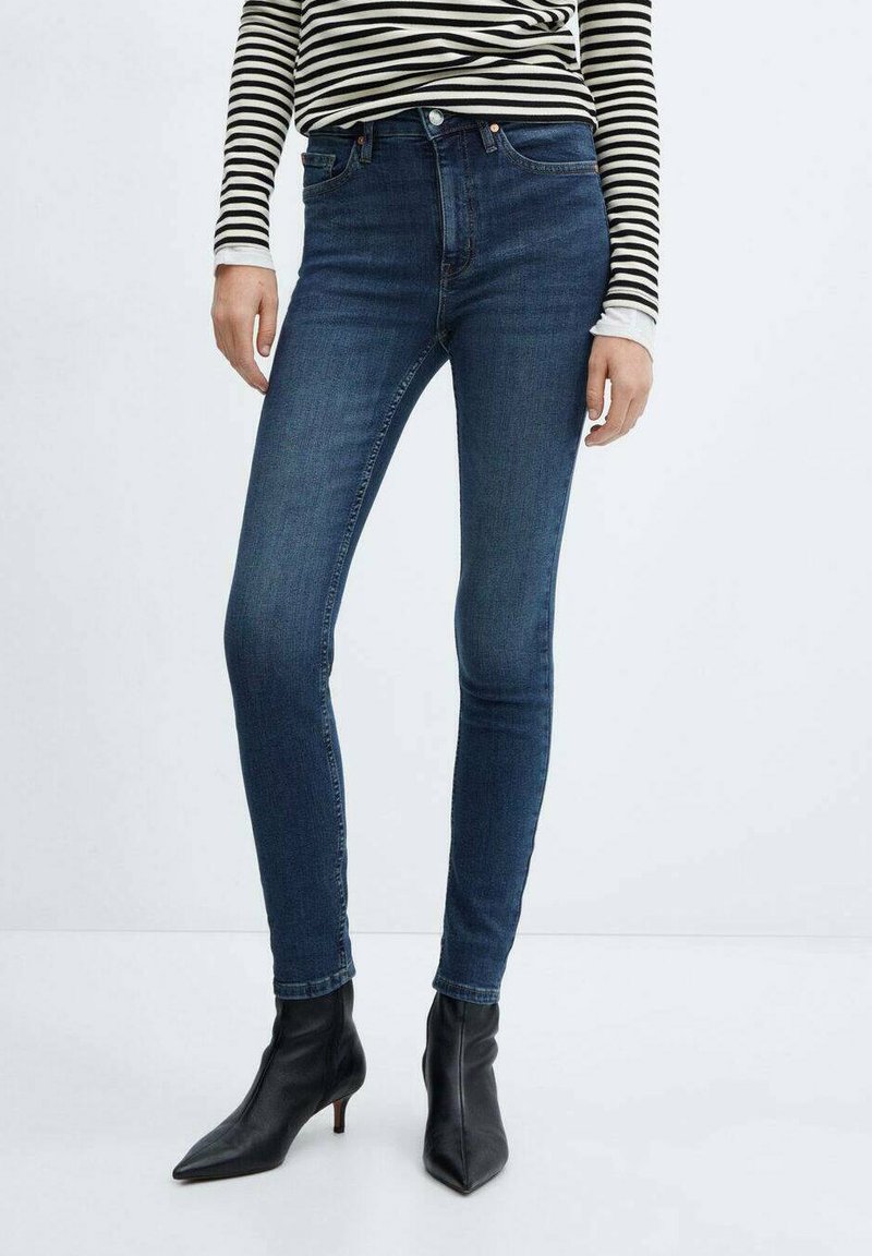Dunkelblaue Skinny-Jeans mit glatter Textur, hoher Taille und minimalen Nähdetails. Kombiniert mit spitzen schwarzen Stiefeletten.
