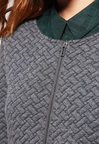 Grå quilted jakke med lynlås og et tekstureret diamantmønster; båret over en mørkegrøn skjorte med krave og ribbet tekstur.